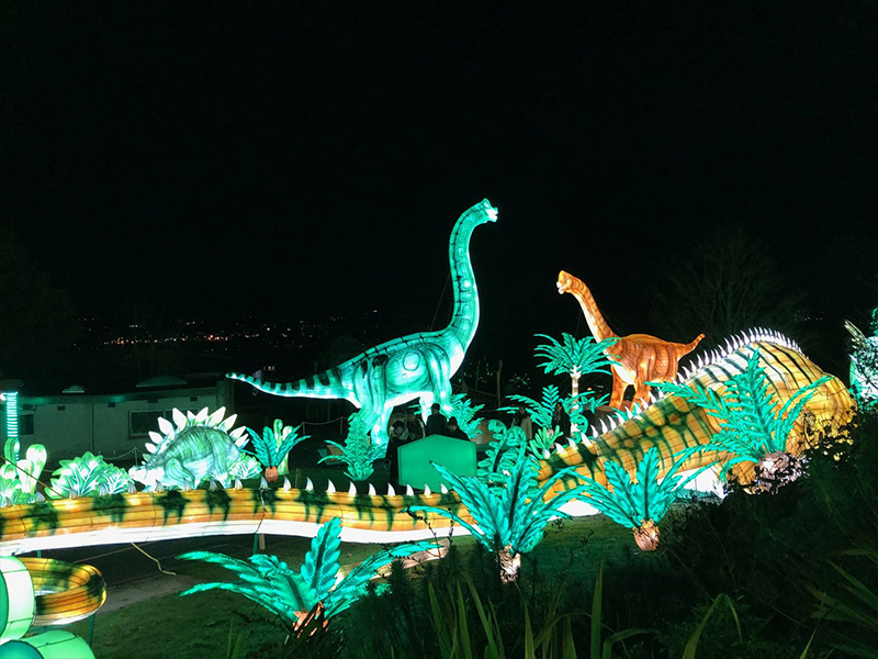 Illuminated sauropod dinosaur lanterns in prehistoric valley scene at night. Long-neck Brachiosaurus figures glow in amber green among palm trees and cycads. Traditional Zigong silk lantern craftsmanship creates immersive Jurassic landscape.