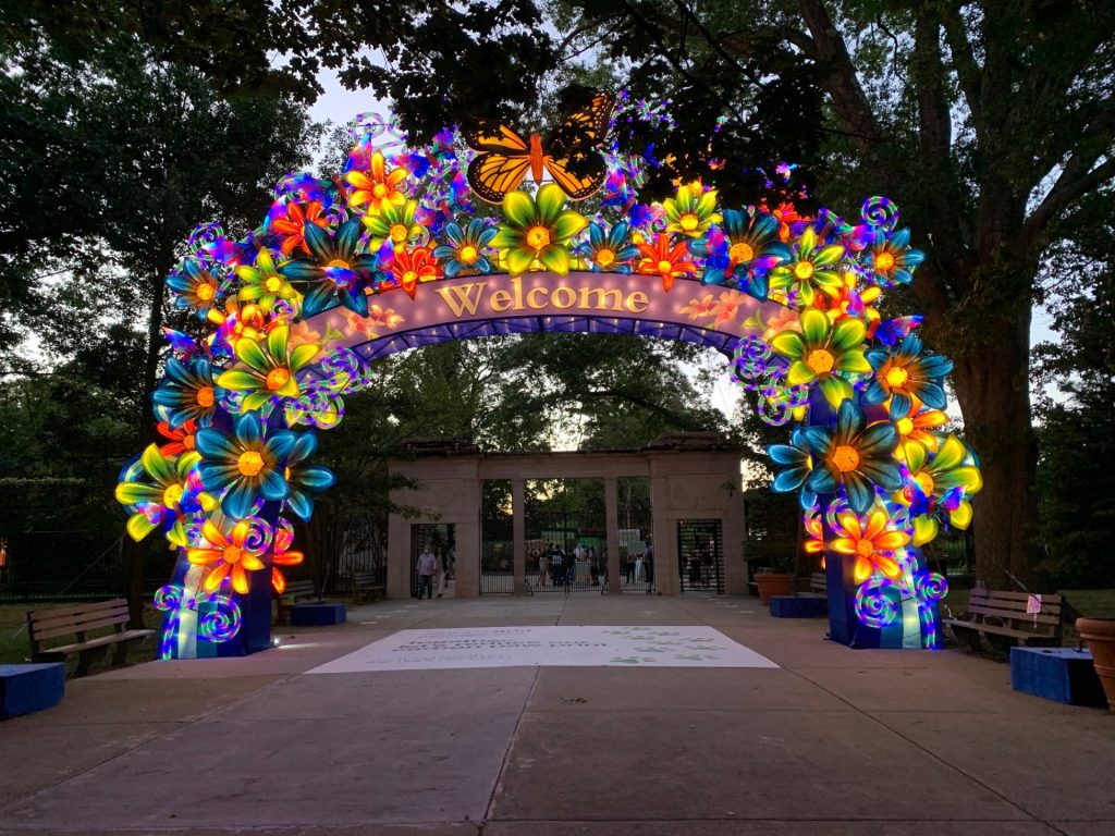 Spectacular illuminated welcome arch at Boston Franklin Park Zoo lantern festival. Rainbow-colored flower blossoms in blue, yellow, orange and pink with monarch butterfly on top. 'Welcome' sign glows warmly as families enter the magical evening experience