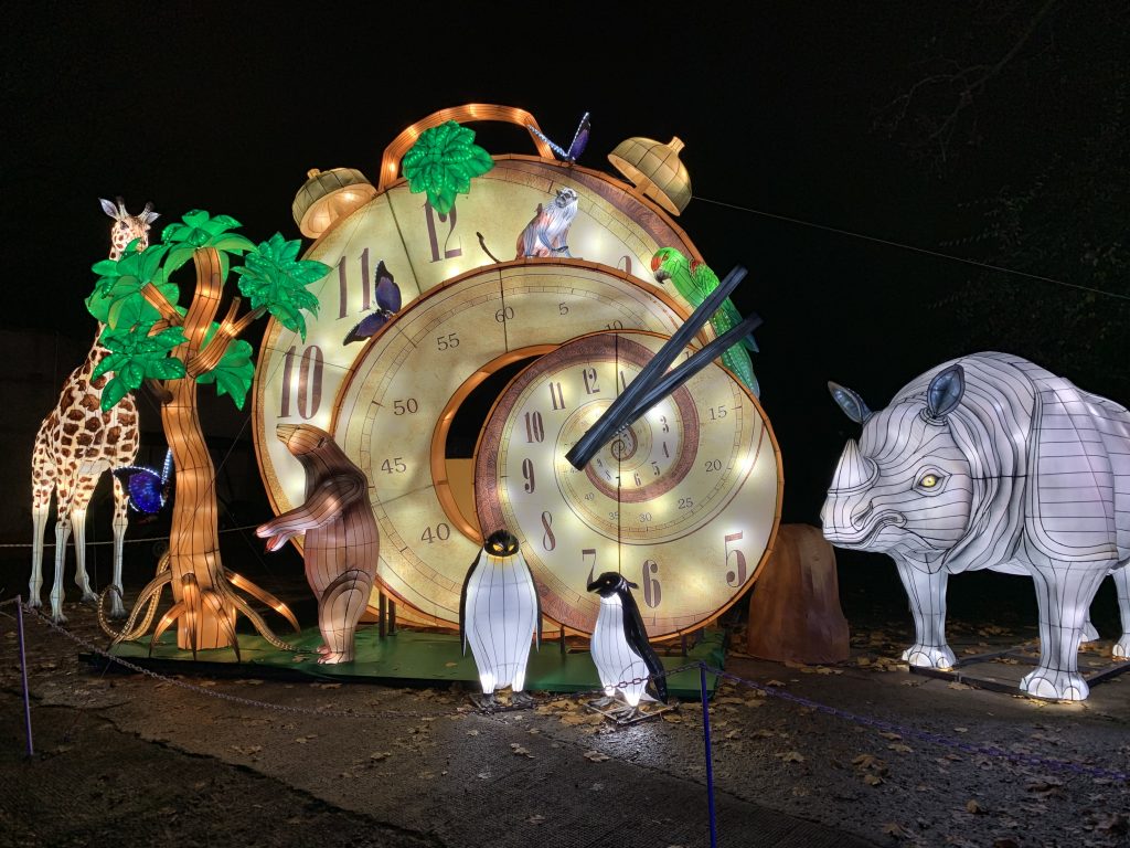 Creative 'Forest Time' lantern installation at Dublin Zoo Wild Lights. Giant golden pocket watch surrounded by animals from different time zones: giraffe, bear, penguins, and rhino. Innovative concept combining Chinese lantern craftsmanship with educational wildlife themes.