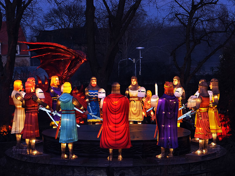 Medieval knights lantern installation at Montréal Botanical Garden Chinese Lantern Festival. Colorful knights in red, blue, purple and gold robes gather around round table with swords drawn, facing dramatic red dragon. Creative fusion of European Arthurian legend and Chinese dragon imagery