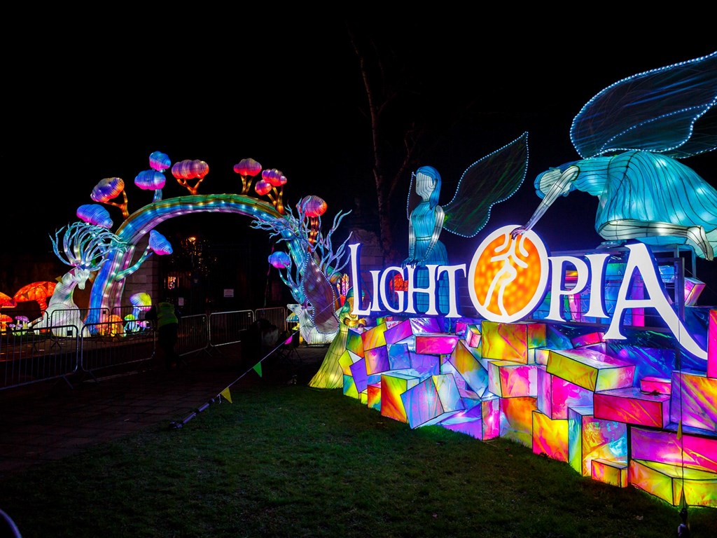 Spectacular Lightopia Manchester festival entrance with giant illuminated logo sign on crystalline rainbow blocks. Fantasy mushroom forest archway and winged ethereal figures create magical welcome. Chinese lantern craftsmanship meets European fantasy design.