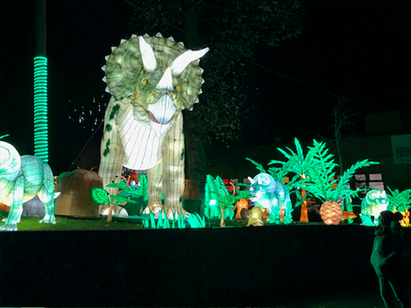Green Triceratops lantern surrounded by colorful illuminated flowers. Detailed horn structure and beak profile demonstrate Zigong lantern makers' anatomical precision. Playful prehistoric garden scene.
