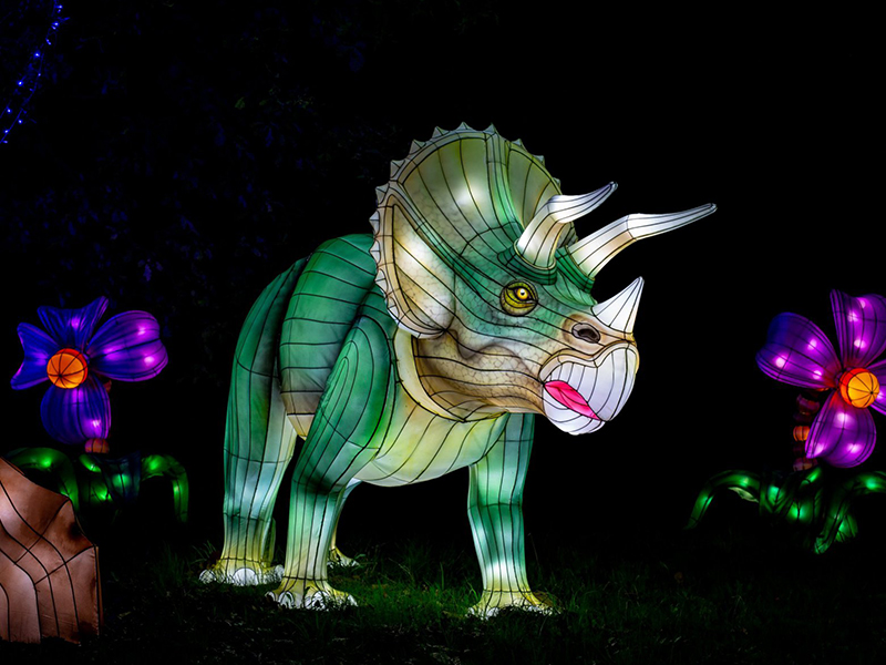 Green Triceratops lantern surrounded by colorful illuminated flowers. Detailed horn structure and beak profile demonstrate Zigong lantern makers' anatomical precision. Playful prehistoric garden scene.