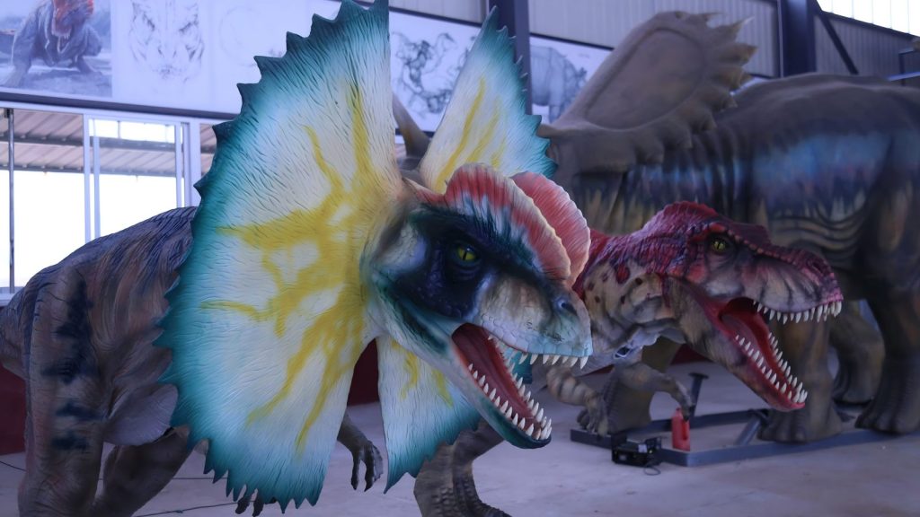 Close-up detail of animatronic Dilophosaurus with expanded neck frill and Spinosaurus showing lifelike eyes, teeth, and textured silicone skin finish.