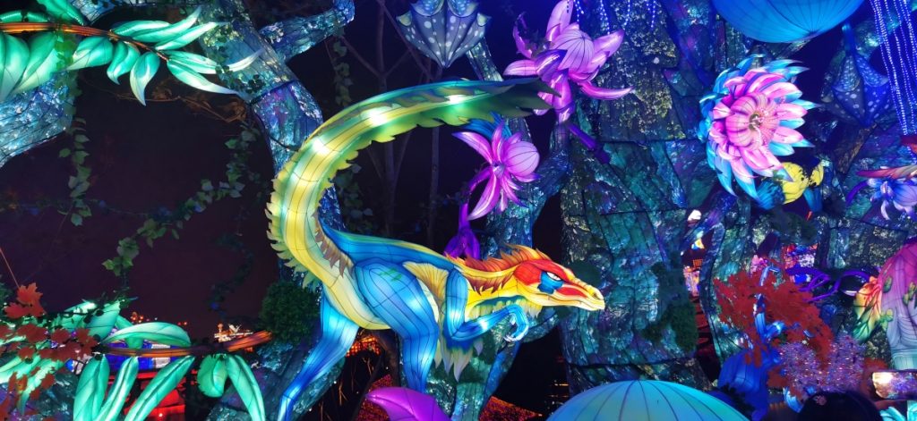 Vibrant dinosaur lantern sculpture with blue-yellow gradient body surrounded by colorful fantasy jungle flowers and glowing tree lanterns — innovative animatronic-style design for themed festivals