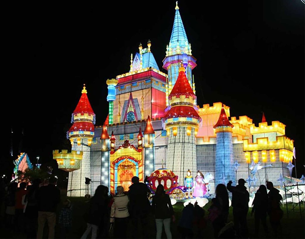 Giant illuminated lantern castle with red and blue towers for Halloween light festival, featuring carriage and princess figures