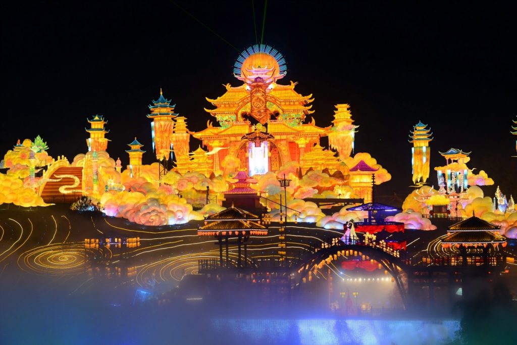 Massive golden palace-themed lantern installation from Zigong lantern festival with multi-tiered pagodas, cloud formations, mist effects, and water reflections creating an immersive celestial wonderland