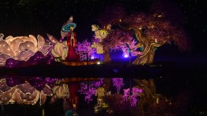 Panoramic Chinese lantern festival display with illuminated animal lanterns, archways, and trees reflected on a night lake