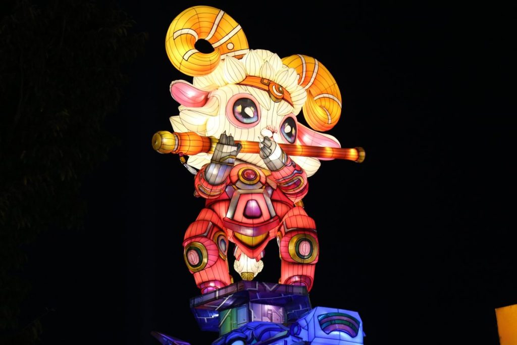 Hand-painted cartoon mechanical sheep lantern holding a glowing golden flute, displayed at night as a zodiac mascot for lantern festivals and theme parks
