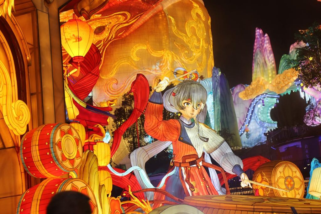 Illuminated Chinese drumming warrior lantern display with a female drummer in traditional costume surrounded by red and gold ceremonial drums