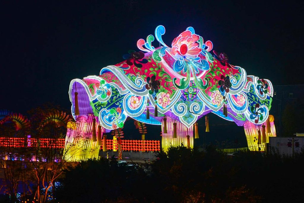Vibrant pink, green, and blue decorative lantern archway with peony flower patterns and tassel decorations, viewed from outside the festival grounds at night
