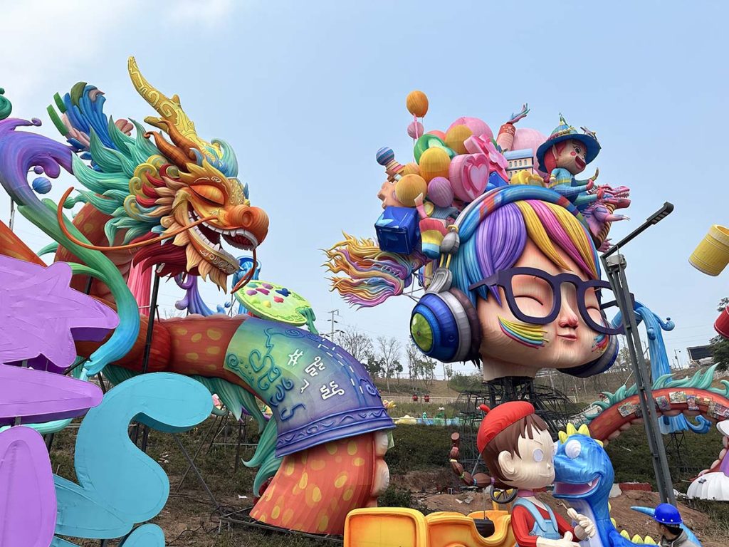 Giant cartoon character lanterns and a multi-colored dragon-phoenix figure during daytime on-site installation, with internal steel scaffolding, support poles, and a worker's hard hat visible