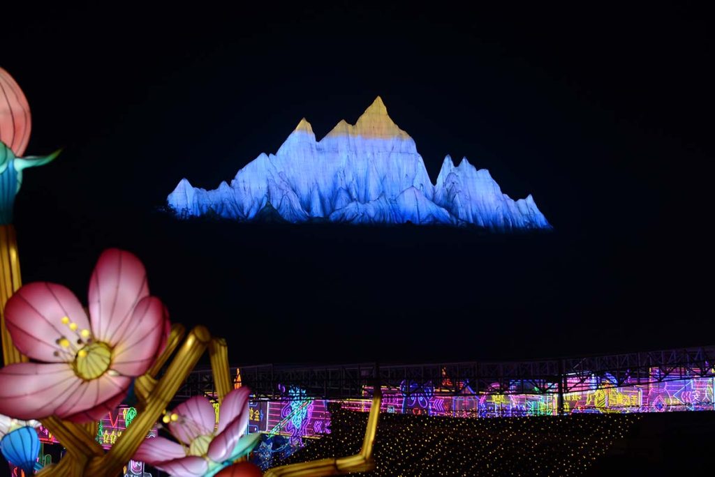 Towering blue-and-white mountain-shaped lantern glowing at night with cherry blossom lanterns in foreground, designed as a panorama-style festival centerpiece viewed from distance