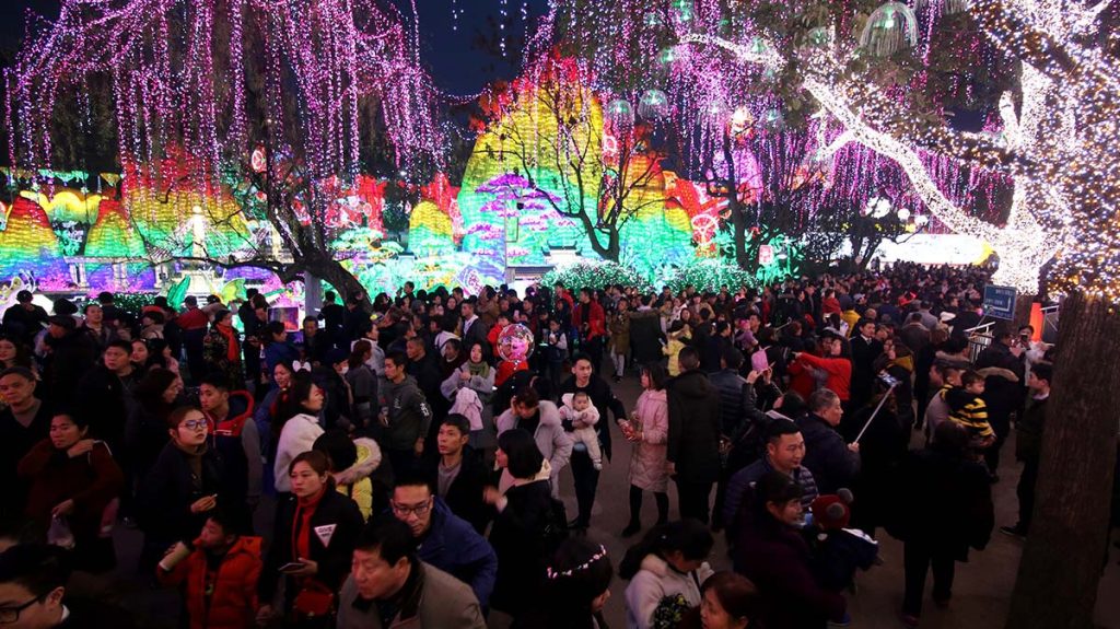 Tens of thousands of visitors at a Chinese lantern festival with pink and white LED-lit weeping willows and colorful mountain-themed lantern displays.