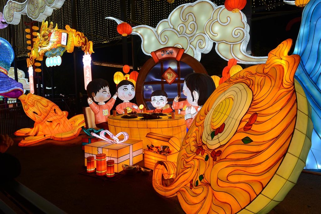 Illuminated Chinese lantern sculpture depicting a Mid-Autumn family reunion dinner with figurines gathered around a round mooncake table framed by auspicious clouds and red lanterns.