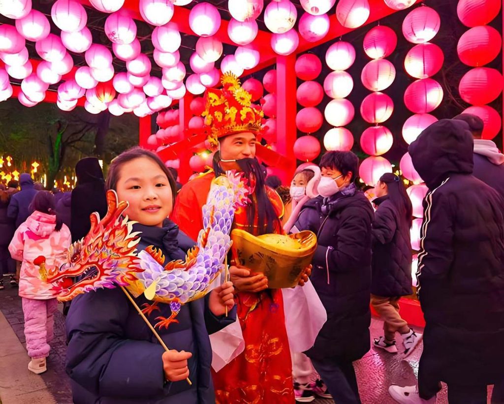 Child posing with a hand-held dragon lantern at a Chinese lantern viewing event, alongside a costumed Caishen God of Wealth performer, under a tunnel of pink and red lanterns.