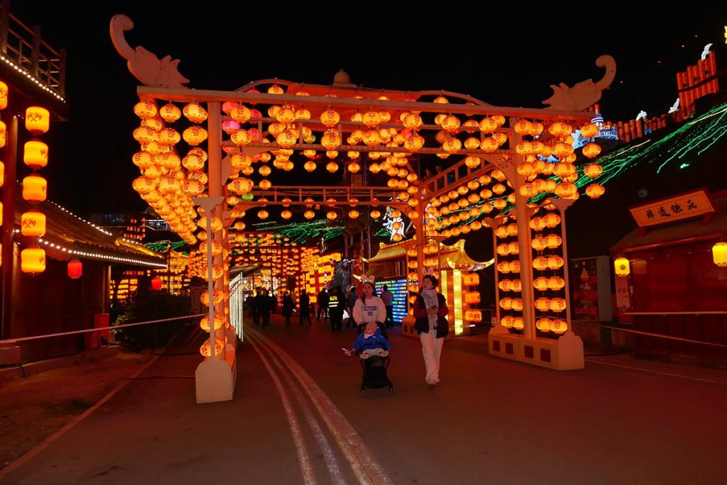Grand lantern tunnel entrance at a Mid-Autumn Festival venue with rows of red and amber lanterns hanging in a wooden archway and mountainside lantern displays in the background.