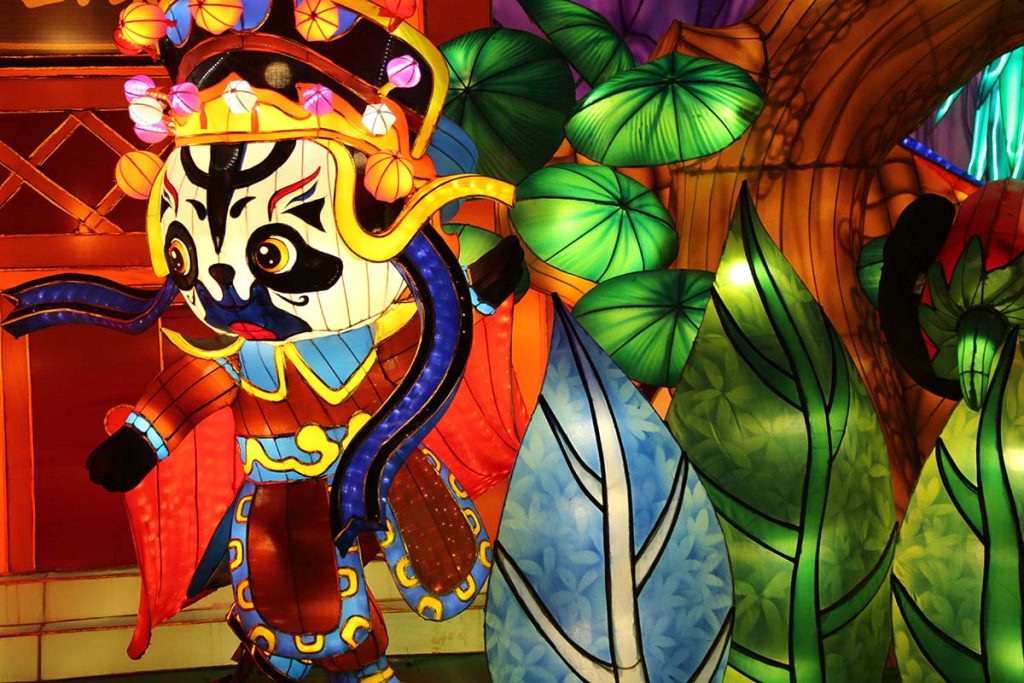 Illuminated Peking opera character lantern with traditional lianpu face painting, ornate pom-pom headdress, blue flowing ribbons, and green lotus-leaf backdrop at a Chinese cultural festival