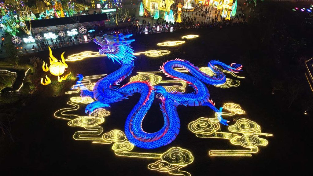 Aerial view of giant programmable RGB Chinese dragon lantern glowing in deep blue and purple color in a festival park at night