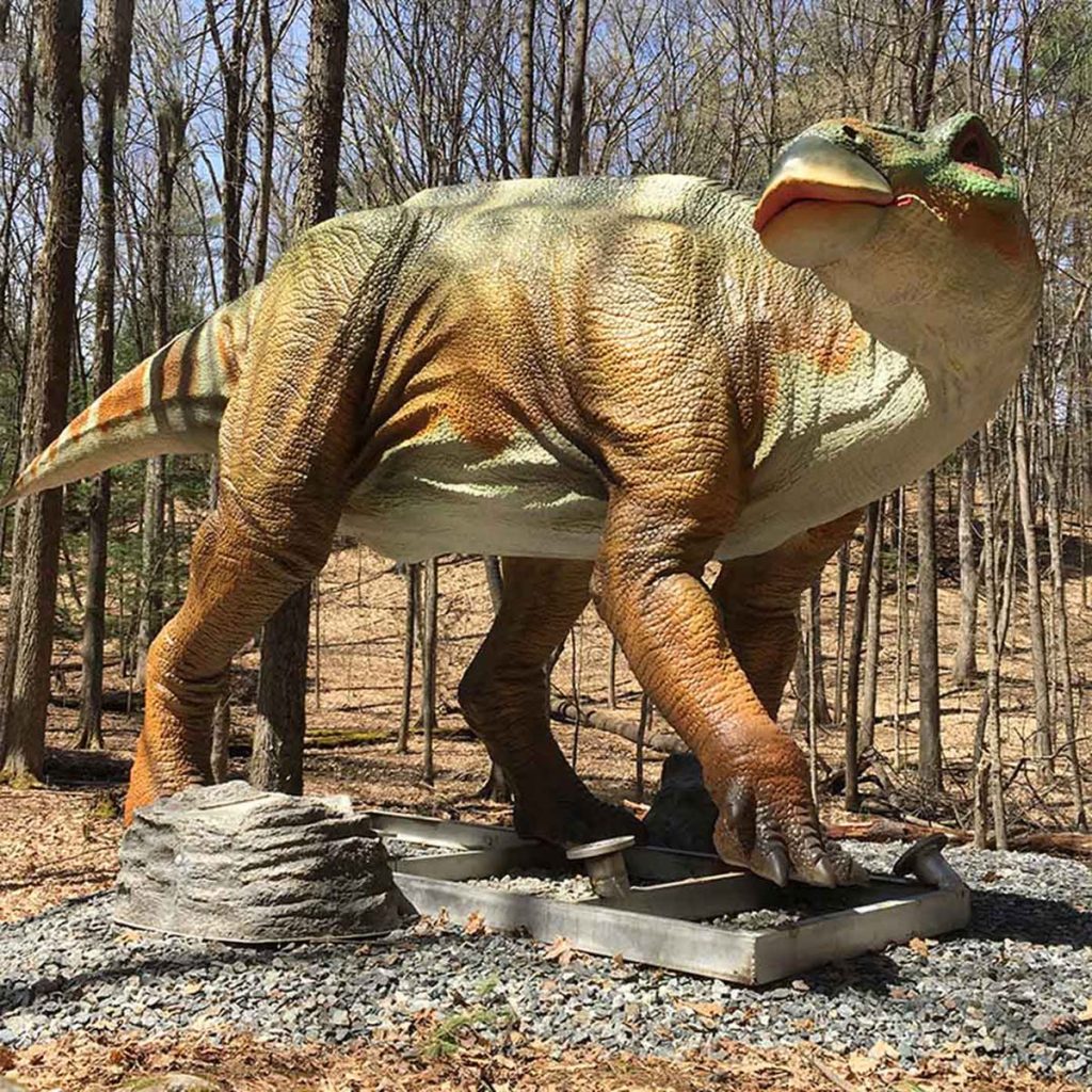 Life-size animatronic Iguanodon (ornithopod dinosaur) in a walking pose on a display base. Tan and green skin with beaked face, bipedal/quadrupedal stance, and stiff tail. Educational exhibit for families and children.