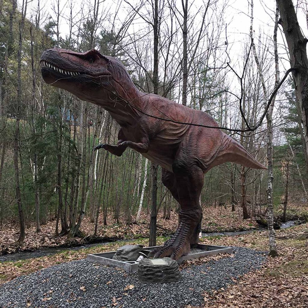 Life-size animatronic T-Rex (Tyrannosaurus rex) in a roaring pose, standing on a base in a wooded outdoor exhibit. Brown silicone skin with textured scales, articulated jaw, and lifelike proportions. Manufactured for theme park centerpieces.
