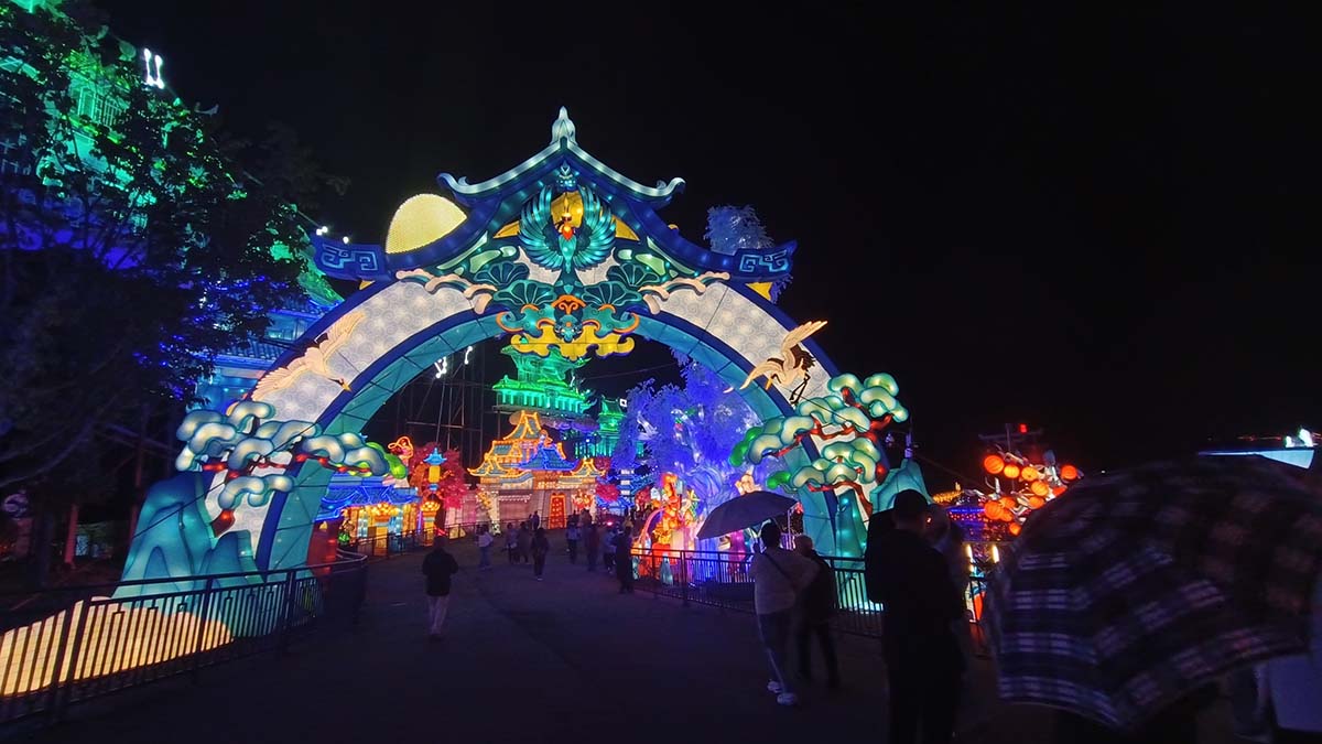 Blue-green phoenix lantern archway with palace lanterns behind and visitors walking under it