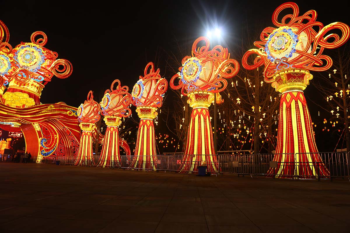 Rows of towering red-and-gold Chinese knot lantern columns line an open plaza at a FestiveLanterns lantern festival, with a red arched gateway in the background