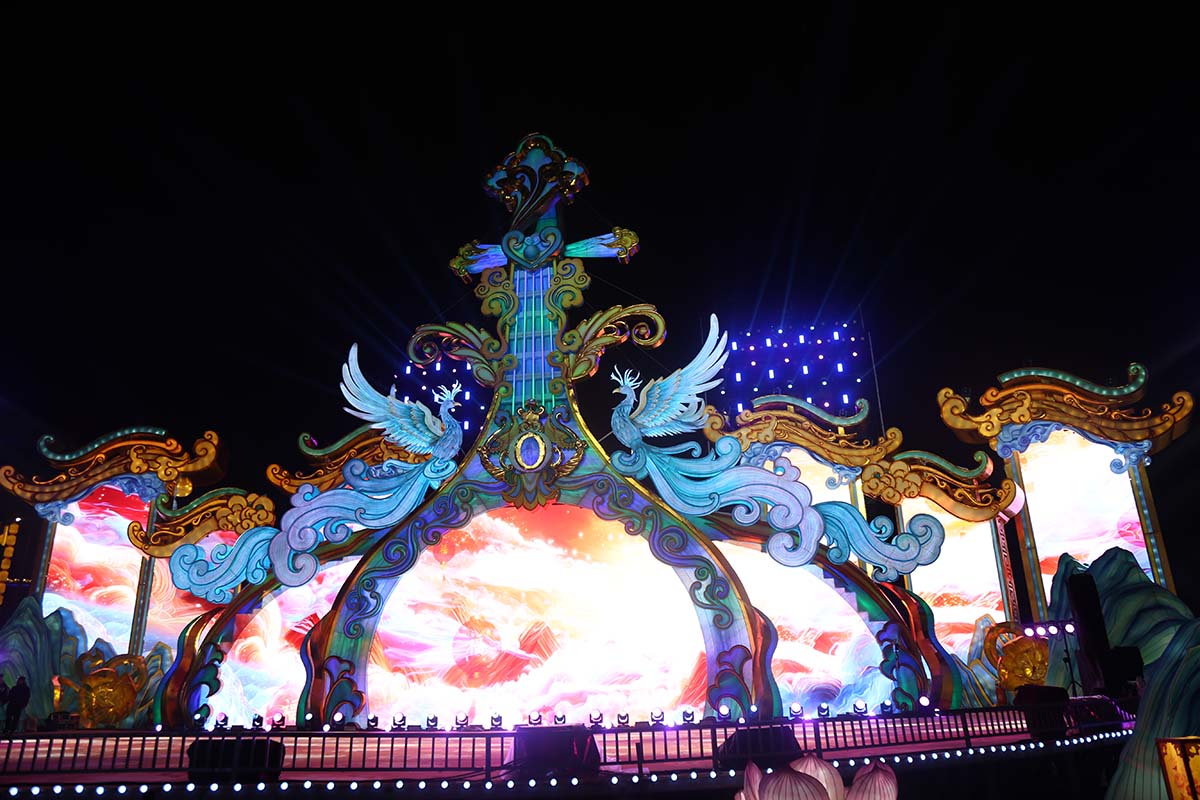A monumental golden phoenix-and-harp lantern centerpiece on a large outdoor stage with LED-lit cloud-screen backdrop at a FestiveLanterns lantern festival