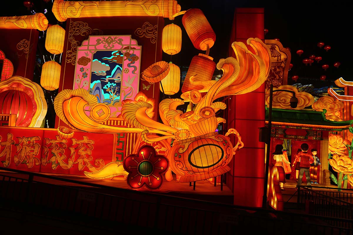 Close-up of a giant red-and-gold traditional Chinese lantern display with a 'Cai Yuan Gun Gun' wishing panel and surrounding red lanterns at a FestiveLanterns festival