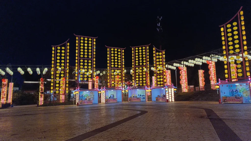 Six Chinese lantern frame display pillars at a city plaza with overhead lantern corridor and cultural display wall at night