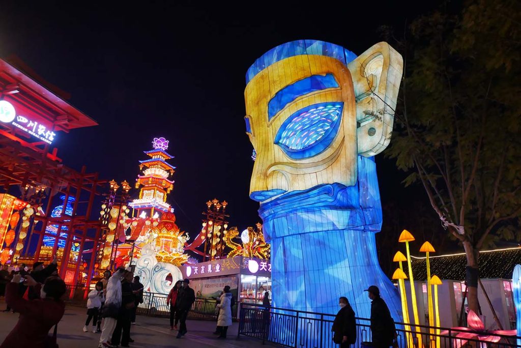Giant Sanxingdui bronze mask lantern display at night with Chinese pagoda lanterns in the background