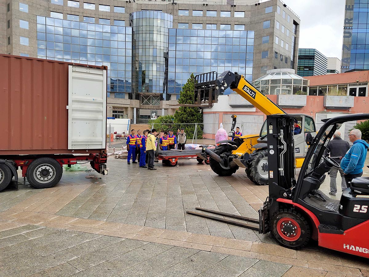 Festival lanterns arriving by shipping container at a European city — workers unloading at venue plaza after sea freight from China