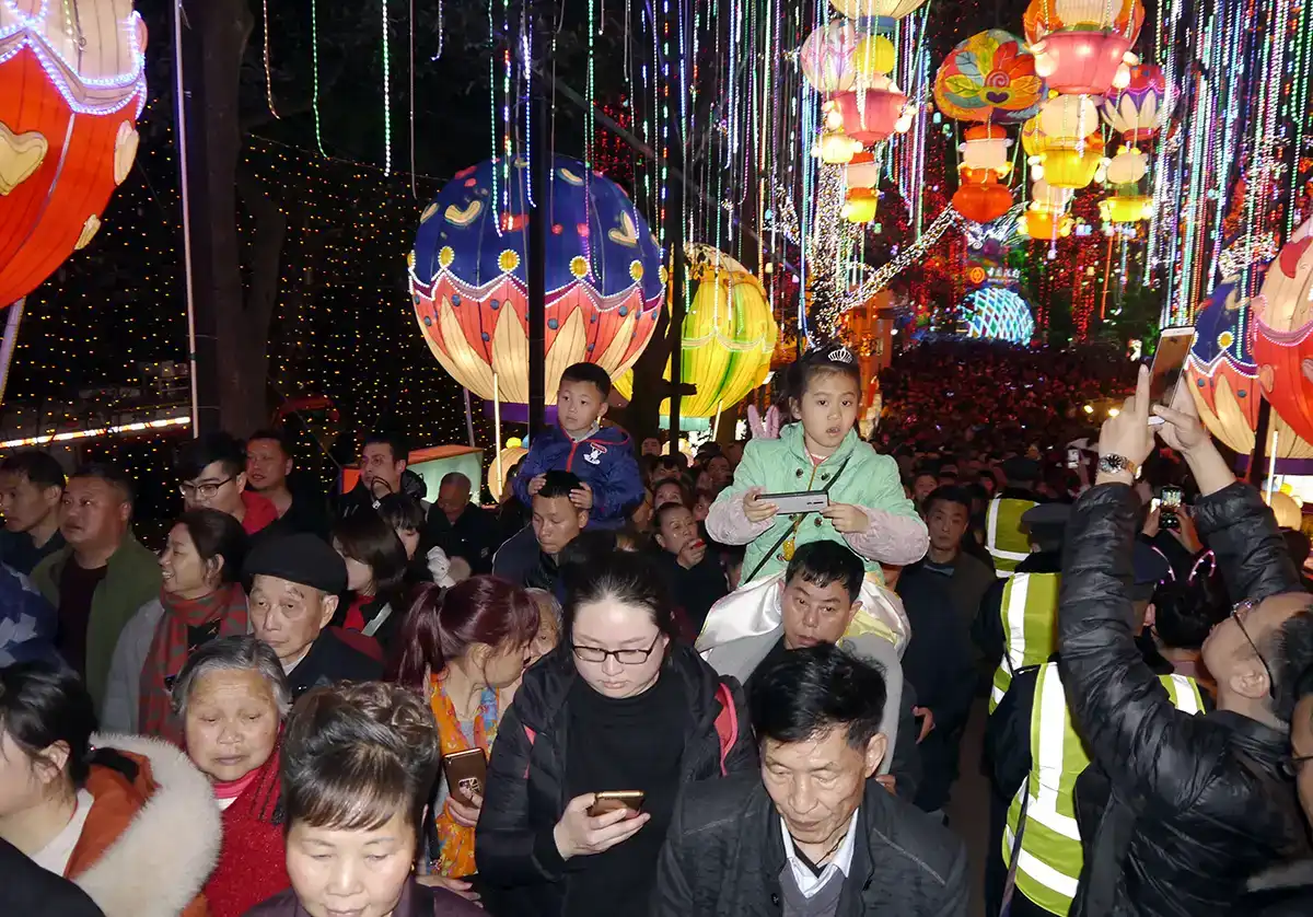 Visitors crowded around a colourful themed lantern display at night — fish and flower shapes hanging above the walkway at a winter lantern festival