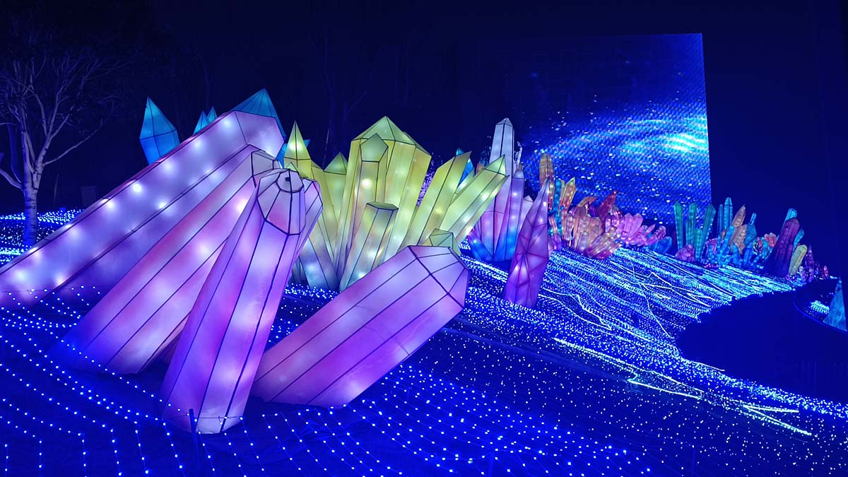 Giant illuminated ice crystal sculptures glowing in purple, green, and gold at a winter lantern festival, with a starry blue backdrop and a sea of blue LED lights on the ground