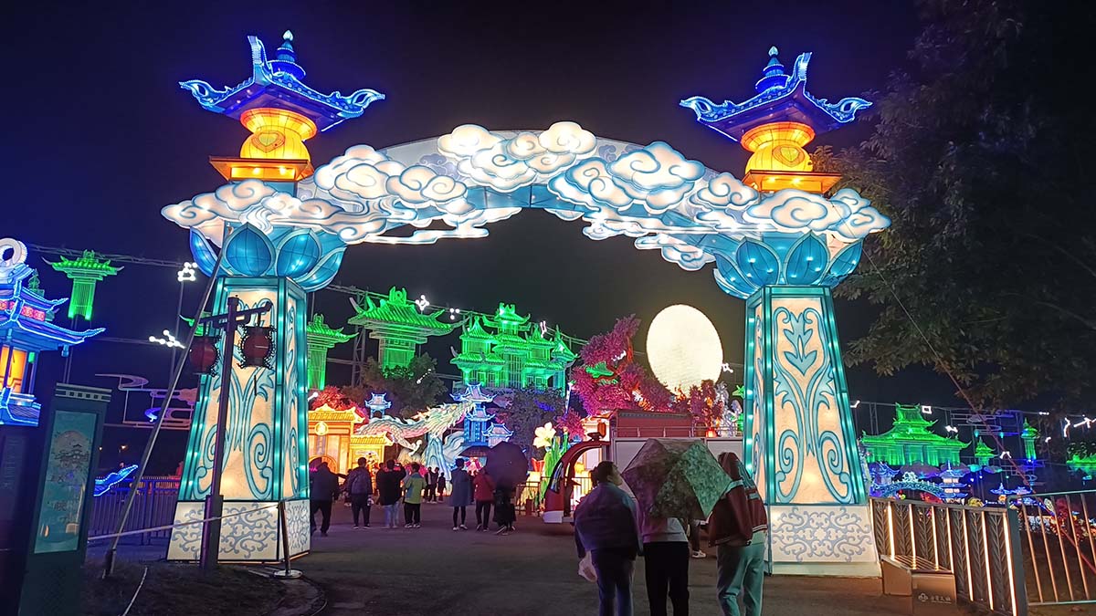 FestiveLanterns winter lantern festival entrance archway with blue auspicious clouds, traditional Chinese pavilions, and a glowing moon above the crowd