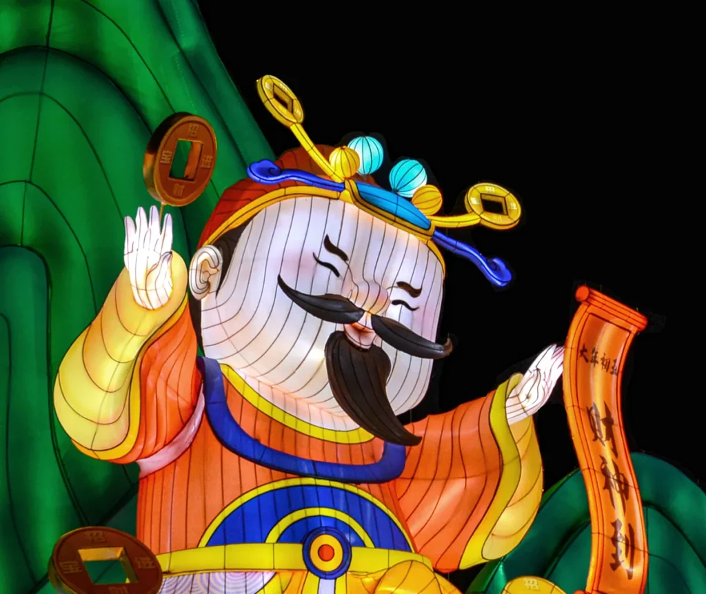 Cartoon god of wealth lantern figure with a raised greeting hand, a red scroll and gold coin lanterns
