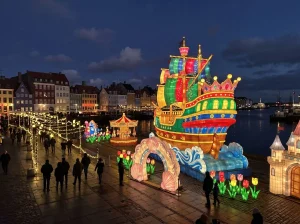 Ship lantern on a harbour promenade — an illuminated sailing vessel with a crown stern, shell archway and carousel beside night-time waterfront buildings