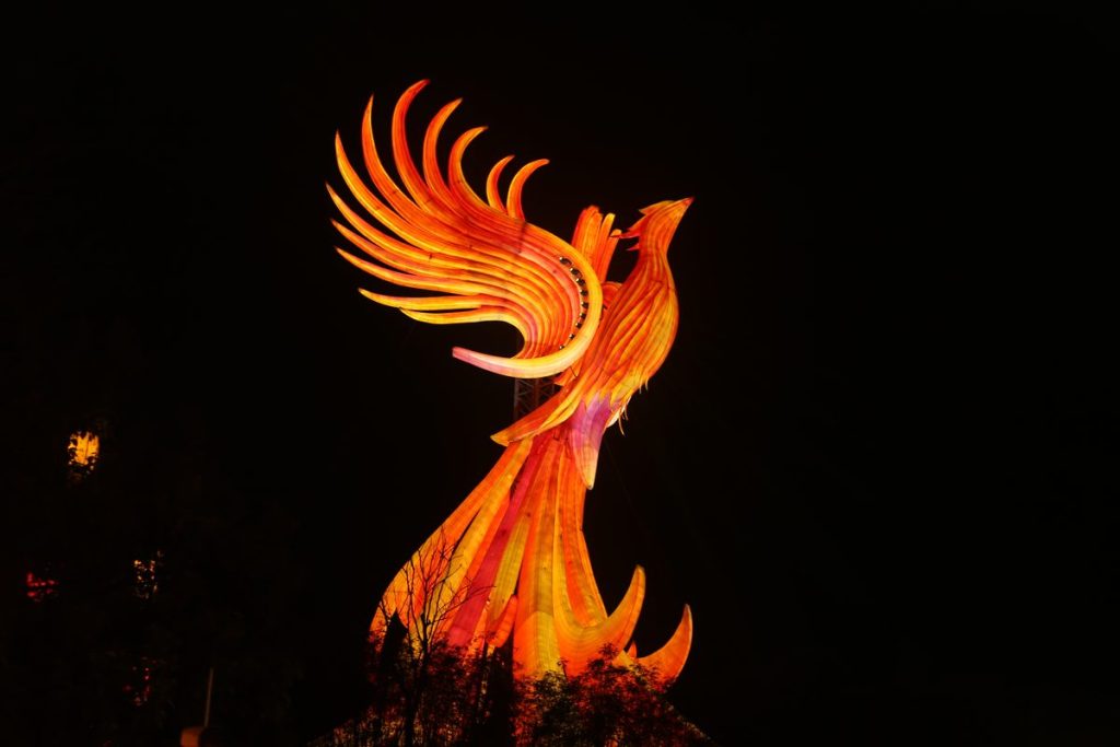 Giant fire phoenix lantern with swept wings and a tail of flame glowing above a rockery base at night